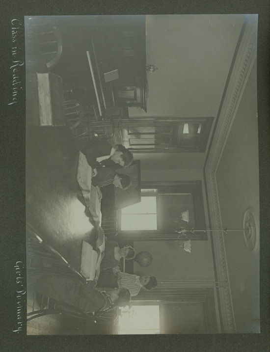 Four girls in dark dresses reading at large table, teacher in light dress standing.