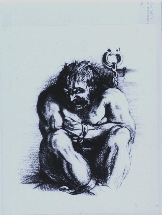 A powerfully built, angry looking man is sitting on the floor, arms folded and knees are drawn up close, his head is turned to the left; his wrists and ankles are in irons which are chained to the wall.