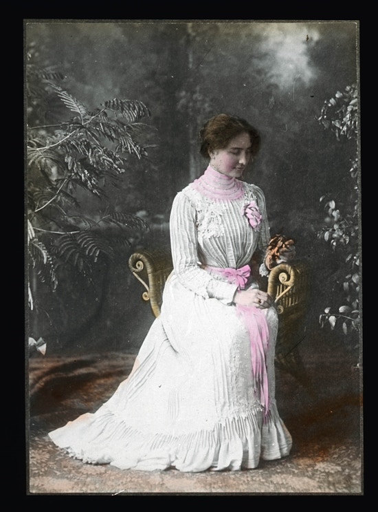 Helen Keller seated wearing a formal white gown with pink ribbon around the waist and holding flowers.