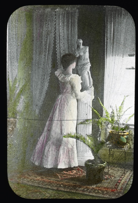 Keller wearing a formal gown, standing to the left of a window, away from camera, with right arm around the base of a statue with potted ferns to the left and right.