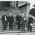 Eight executive committee members of the American Association to Promote Teaching Speech to the Deaf stand on outdoor steps for a black and white photo.