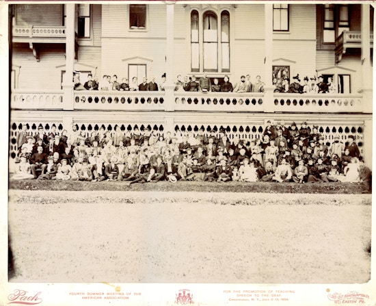 Large group photograph of American Association To Promote Teaching Speech To The Deaf summer meeting attendees wrapped above and below a large white porch in 1894.