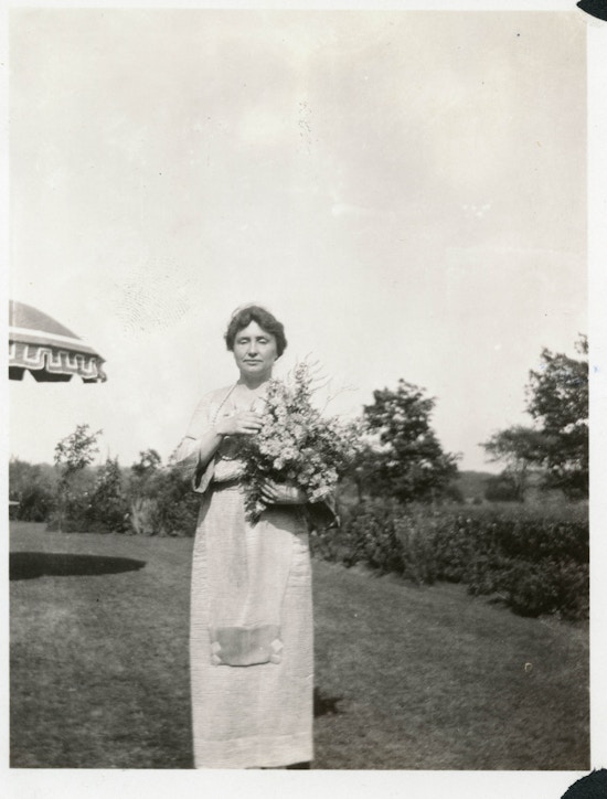 Helen Keller in the garden of her Long Island home (Forest Hills, NY), taken on her birthday by Rebecca Mack. Stands on lawn holding flowers.