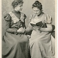 Anne Sullivan (right) finger spelling a book with Helen Keller (left), both seated.