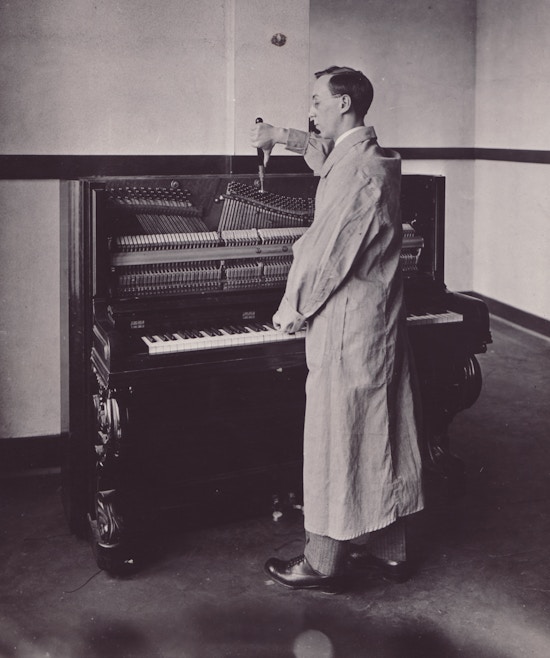 Man in white lab coat stands in profile at piano, working with tuning instruments.