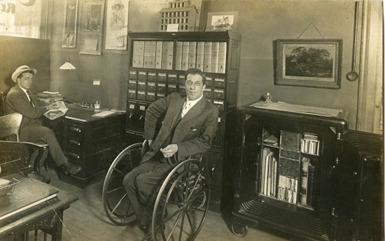 Man using wheelchair in an office, another man at a desk.