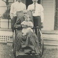 Two Men, Baby, And Elderly Woman In Wheelchair.  Next to building with porch.