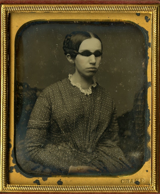 Portrait of Laura Bridgman as a young woman wearing a patterned dress with lace collar. Her hair is braided and pinned up with a part down the middle. The Daguerreotype photograph is housed in a small leather case with a patterned red velvet lining surrounded by a gold mat board and embossing.
