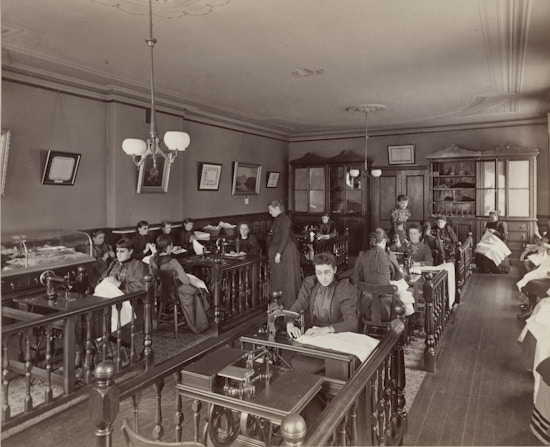 Girl's sewing class from upper grammar at the the Perkins Institution and Massachusetts School for the Blind in South Boston. The sewing room has eight or more sewing machine tables in two rows with railings sectioning off the work stations. Students sit on benches along the walls with sewing in their laps.