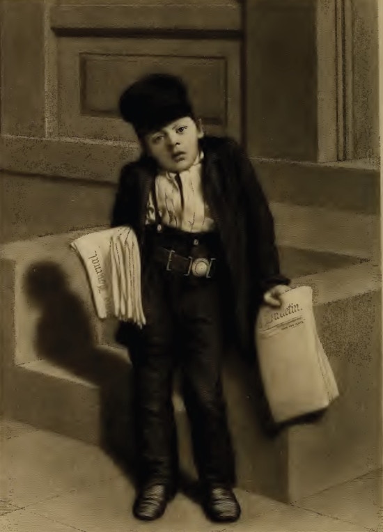 A boy wearing high-waist pants, an over-sized jacket, and a hat holds newspapers in both hands.