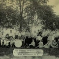 Photograph of large group of musicians posing with their instruments under a tree