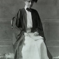 Helen Keller sits in a straightback wooden chair wearing cap and gown.