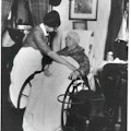 Elderly woman in wheelchair, with blanket over her legs, is tended by nurse.
