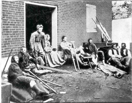 Soldiers sit or lie on the ground in an outdoor courtyard. A nurse helps a soldier on crutches out the door.