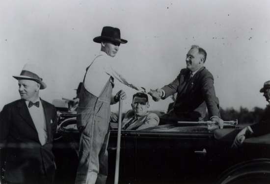 President Roosevelt sits up on the back of a car seat and shakes hands with a farmer in overalls standing next to the car.