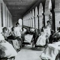 Seven women sit in large wheelchairs on the porch of a large Victorian house at a sanitarium. Each is covered in a blanket and looks passively at the camera. Six attendants and two children stand behind and amongst them.
