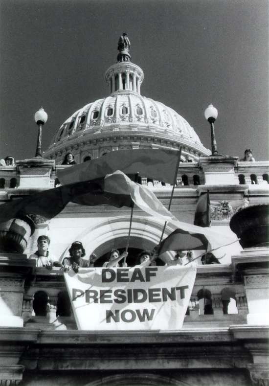 Seven deaf students protest. They wave flags and stand behind a banner that reads "Deaf President Now".