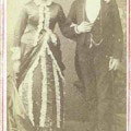 A portrait of a couple standing, the woman in a dress with ruffles and the man in a suit.
