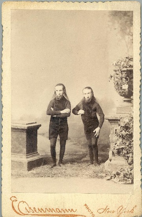 Two small-statured men with long hair and beards stand between two pedestals.