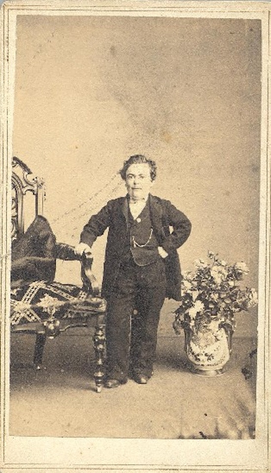 A short-statured man in formal dress stands with one hand on a chair.