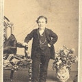A short-statured man in formal dress stands with one hand on a chair.