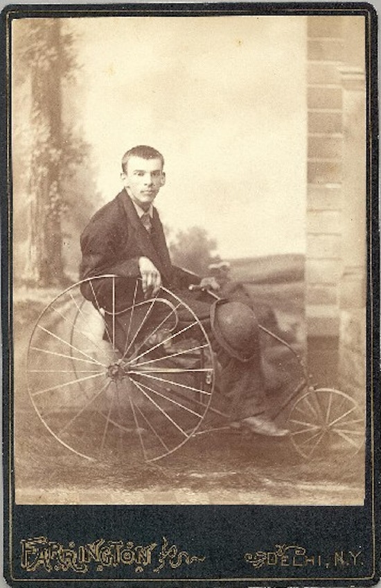 A young man, with hat on his knee, sits in a wheelchair with metal wheels.