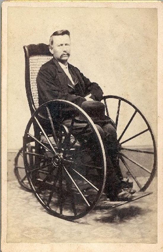 A man, with a hat on his lap, sits in a wheelchair.