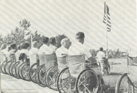 A row of men in wheelchairs before an American flag.