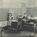 A teacher and a girl stand at a blackboard.  Four boys sit at their desks.