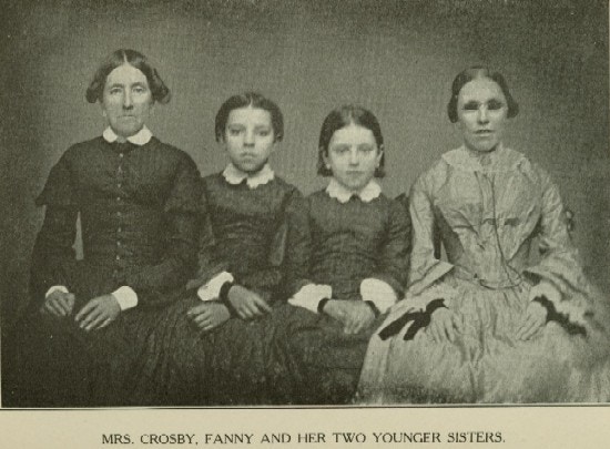 A photographic portrait of a women in a plain black dress with her three daughetrs, one wearing dark glasses.