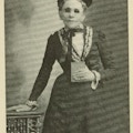 shows a woman in a black dress with white lace and a black bow at the neck holding a small book in her left hand and her right hand on the top of a short table to her right