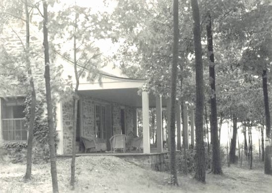Porch, with wicker furniture, seen through trees. Ivy covers part of house.