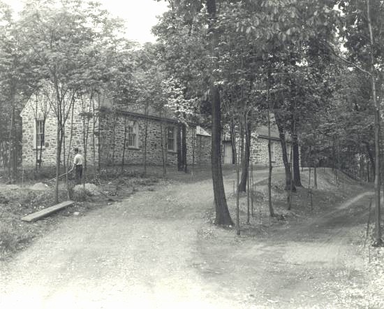Stone house seen through trees, with a dirt driveway making a switchback up a hill. A man works with a shovel next to the house.
