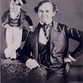 P.T Barnum sits in a chair.  Tom Thumb, with hand on Barnum's shoulder and in uniform, stands on table.