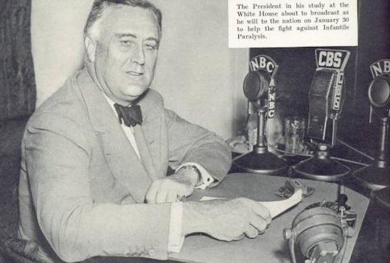 FDR sits at desk with CBS and NBC radio microphones.