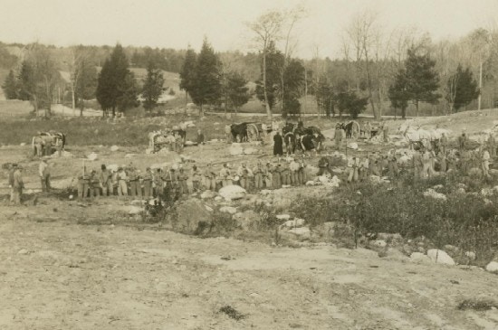 A row of men, with horses and wagons in background, work clearing the ground of debris.
