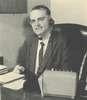 A man in a suit sits at a desk and wears a telephone headset.