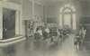 Children sit at their desks with books open. A crutch leans in the corner.  Two steps access a hall.