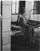 A partially clothed man sitting on a bed next to a barred window.
