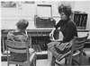 A boy, in a chair marked Fred, sits next to a woman and a record player.