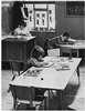 A teacher at her desk and two children at tables.