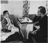 A boy sits on a bed with a tray of food and speaks with a woman.