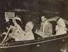 Franklin Roosevelt drives a car as King George speaks to him from the back seat.  Roosevelt is seated next Queen Elizabeth.