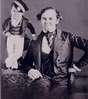 P.T Barnum sits in a chair.  Tom Thumb, with hand on Barnum's shoulder and in uniform, stands on table.