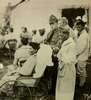 Photograph of injured Native American soldiers, dressed in robes, at what appears to be a military field hospital.  The soldiers smoke.  In the foreground, one soldier uses a heavy wheelchair.