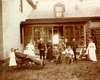 A family poses in front of the porch of a house.  An elderly man lies in a wheel chair.