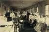 Photograph of ward in military hospital, including veterans, nurses, and doctors.  Several men use wheelchairs.