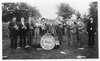 A nine-piece brass band stands in a field in front of trees holding their instruments.  They stand around a large drum on which is printed State Insane Asylum Band, Topeka, KS.