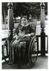 A young girl, holding a book, sits in a wheelchair on a porch.