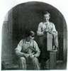 Two boys working as cobblers. One files the sole of a shoe. The other holds a hammer and last upright and smiles.